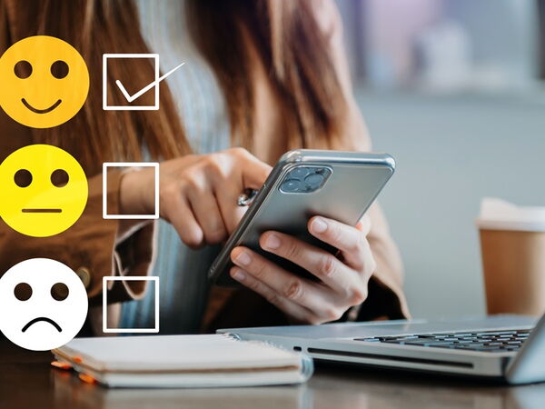Eine Frau sitzt am Schreibtisch mit einem Smartphne in der Hand, auf dem Foto sind 3 Smilies von gut bis schlecht, wobei gut angehakt ist